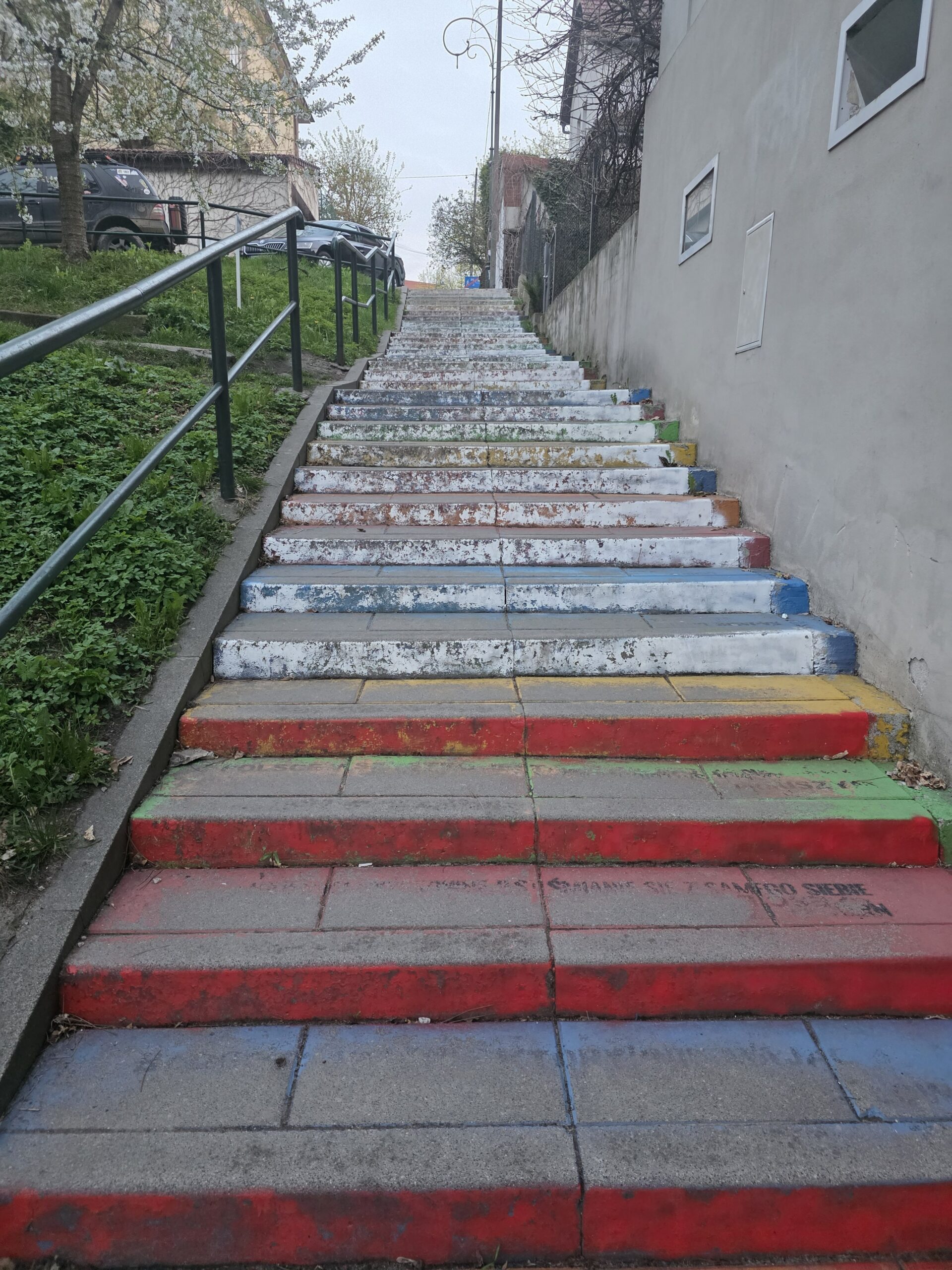 Kolorowe schody w Krakowie w dzielnicy Podgórze, pomalowane na różne barwy, prowadzące w górę między budynkami. Stopnie mają wyraźne ślady zużycia i wyblakłe fragmenty farby, co pokazuje działanie czasu i warunków atmosferycznych. Schody znajdują się w zielonej, spokojnej części miasta, otoczonej miejską zabudową i roślinnością, tworząc kontrast między sztuką uliczną a codzienną przestrzenią Krakowa.